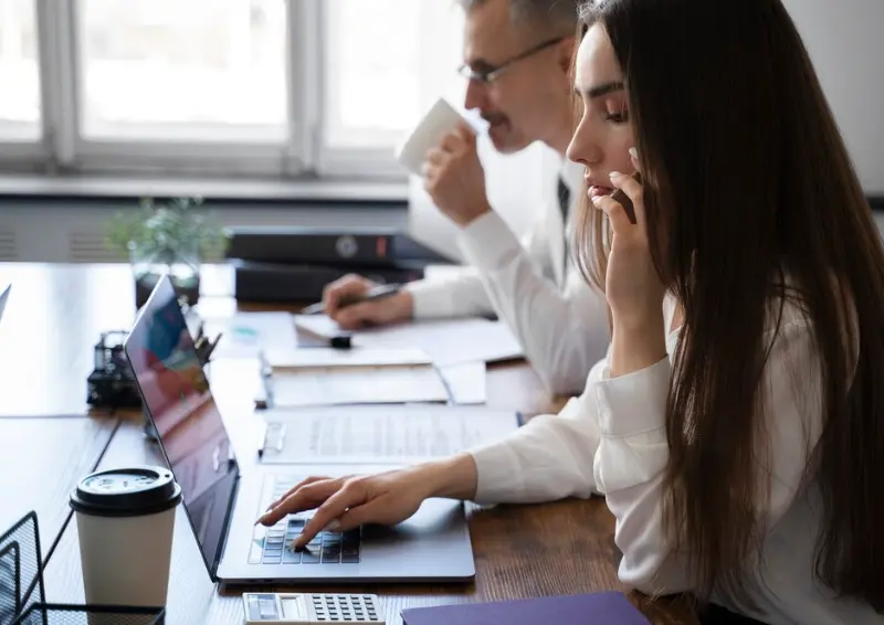 Young businesswoman multitasks on phone and laptop while colleague reviews documents and sips coffee in bright office